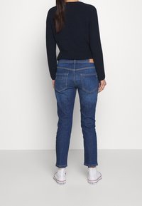 Slim fit blå denimjeans med fem fickor och en ljusbrun läderlapp på bakre midjebandet. Modellen har på sig vita sneakers.