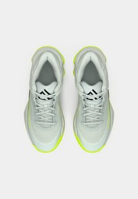 Par af lysegrå atletiske sneakers med sort logo på tungen og neongule såler, vist fra topvinkel på en hvid baggrund.