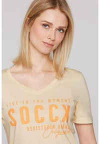 Camiseta de algodón amarilla clara con mangas cortas y escote en V, que presenta un texto en negrita de color naranja, incluyendo "SOC CX" y acentos en cursiva.