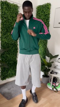 En mann iført en grønn og rosa Adidas-jakke, lysegrå shorts, hvite sokker og svarte loafers står innendørs nær planter og et lite bord.