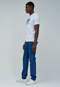 Camiseta blanca de manga corta con un gráfico de "SPLASH JEANS" en azul y negro, combinada con jeans de mezclilla azul y zapatillas blancas.