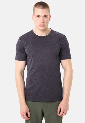 Junger Mann mit kurzen Haaren, der ein dunkelgraues, eng anliegendes T-Shirt und olivgrüne Hosen trägt, vor einem schlichten hellen Hintergrund stehend.