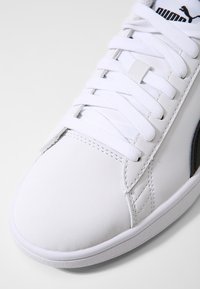 Vit syntetisk sneaker med en slät yta, som har platta vita snören, en vadderad tunga och en svart accentrand på sidan.