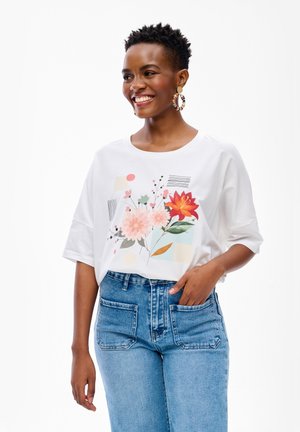 Uśmiechnięta kobieta o krótkich, kręconych włosach, nosząca duże kołowe kolczyki, białą bluzkę w kwiatowy wzór oraz niebieskie dżinsy z wysokim stanem i przednimi naszywanymi kieszeniami.