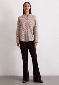 Blouse à manches longues et col, avec un fond beige et un motif à pois marron. Associée à un pantalon évasé noir et des chaussures marron.