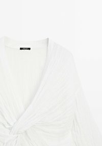 Witte wikkeltop van textuurstof met een knoopdetail in de taille en wijde mouwen. Label zichtbaar op de halslijn.