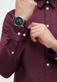 Camisa burdeos abotonada con textura suave y botones blancos, usada mientras se ajusta un reloj de pulsera negro con caja plateada.