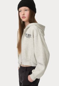 Korte lichtgrijze hoodie met een voorzijde rits en geribbelde tailleband, met zwarte tekst "THE BRONX Sports District N.Y.C." en een beanie.