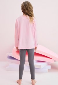 Rosa långärmad tröja och grå leggings, tillverkade av mjukt material. Påfallande detalj: liten regnbågsgrafik på ärmen. Modell vänder bort.