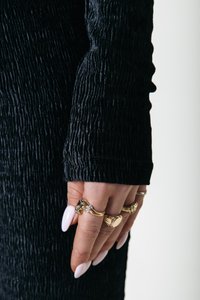 Schwarze, strukturierte Stoffhülle mit Falten, die ein enganliegendes Design aufweist. Im Vordergrund sind zahlreiche goldene Ringe an den Fingern mit langen, weißen Nägeln zu sehen.