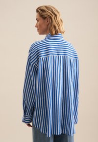 Blue and white striped shirt with a loose fit, button-down collar, long sleeves, and a curved back hem. Made of lightweight fabric.