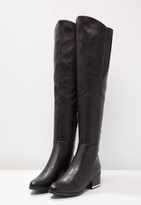 Bottes en cuir noir au-dessus du genou avec une texture lisse, un talon carré et un détail de fermeture éclair sur le côté. Bout légèrement rond et panneau élastique.