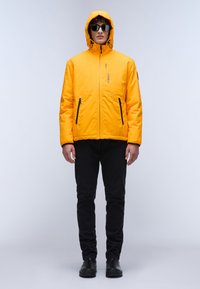 Geel geïsoleerde jas met een capuchon, frontale rits en twee zwarte ritszakken. Draagt over zwarte broek en zwarte schoenen.