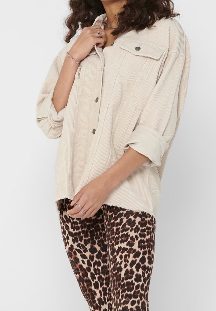 Femme portant une veste en velours côtelé beige avec les manches retroussées et un pantalon imprimé léopard, accessoirisée avec des bagues et un bracelet.