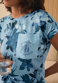 Moteris vilkinti mėlyną gėlių marškinėlį su išsiuvinėta kišene, kurioje parašyta "By Ted Baker", laiko stiklinę vandens.