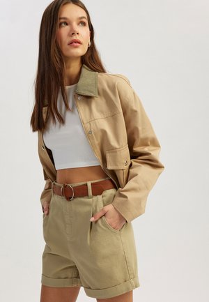 Chaqueta recortada beige con cuello verde, combinada con pantalones cortos khaki de cintura alta y un cinturón marrón con una hebilla circular.