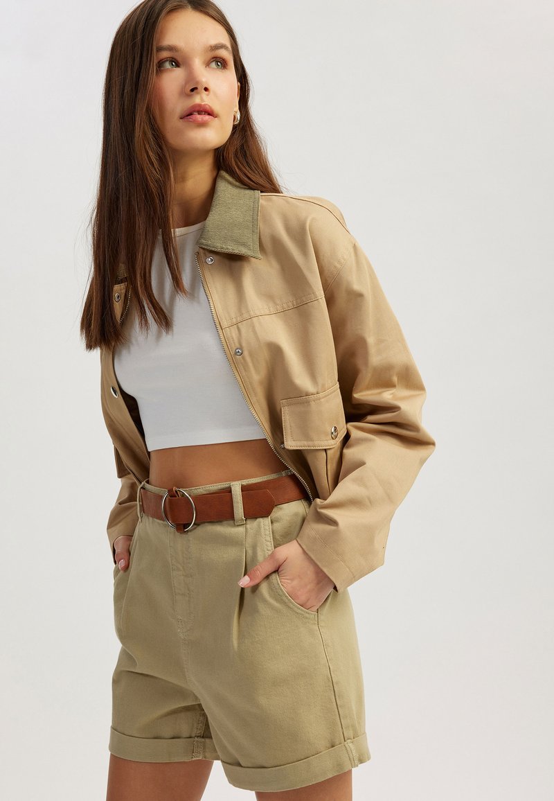 Beige cropped jack met groene kraag, gecombineerd met hoge kaki shorts en een bruine riem met een ronde gesp.