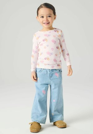 Enfant souriant portant une chemise rose à manches longues avec un motif de ballons, un jean bleu clair à jambes larges avec des patchs de ballons, et des chaussures marron à enfiler.