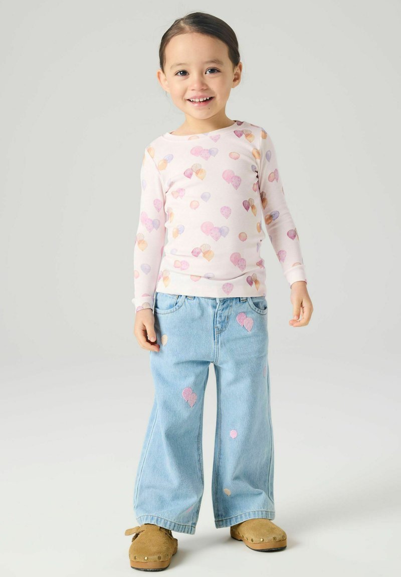 Mosolygó gyermek hosszú ujjú, rózsaszín léggömbmintás ingben, világoskék, bőszárú farmerral, léggömb foltokkal, és barna, könnyen felhúzható cipővel.