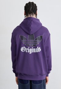 Lilla hettegenser med en stor svart og sølv Adidas-logo på ryggen, med snøring på hetten og ribbestrikkede ermer.