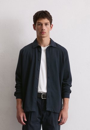 Navy button-up shirt with a spread collar, long sleeves, and a relaxed fit, worn over a white crew neck t-shirt and paired with dark pants.