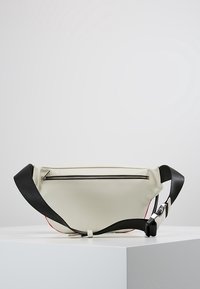 Bolsa de material suave y blanca con una correa negra, que presenta un cierre superior con cremallera y acentos rojos a lo largo de los bordes. Diseño elegante y minimalista.