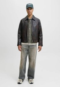 Braune Lederjacke über einem schwarz-weißen Kariertshirt, kombiniert mit hellen, ausgebleichten Jeans und beigen Turnschuhen. Mann steht vor einem weißen Hintergrund.