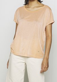 Lyhythihainen persikkavärinen paita, jossa on pehmeä, hohtava pinta ja pyöreä kaula-aukko. Paidassa on pieni metallinen logodetaili helmassa.