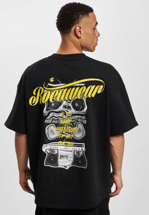 Černé oversized tričko s grafikou hromaděných boomboxů v šedém a žlutém písmu, s nápisem "Rocawear" výrazně umístěným.
