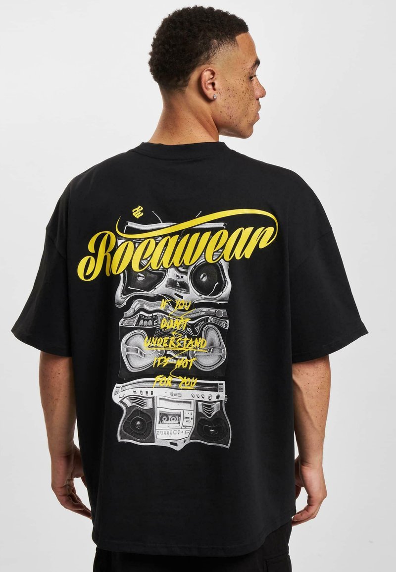 Černé oversized tričko s grafikou hromaděných boomboxů v šedém a žlutém písmu, s nápisem "Rocawear" výrazně umístěným.