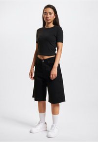 T-shirt court noir, short en denim noir longueur genou avec ourlet effiloché, baskets blanches et chaussettes blanches montantes. Design minimaliste et coupe ajustée.