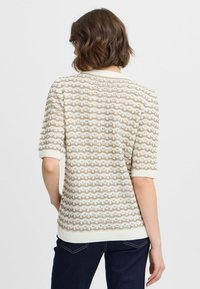 Suéter de punto en crema con un patrón de olas, con mangas cortas y acabados acanalados. Tela ligera y texturizada con sutiles detalles en dorado.