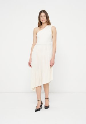 Vrouw draagt een asymmetrische witte geplooide jurk met één schouder, gecombineerd met zwarte hoge hakken met enkelriem, staand tegen een witte achtergrond.