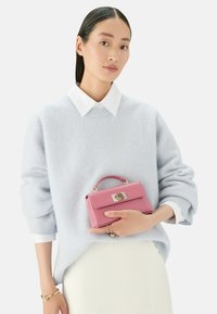 Světlomodrý oversized svetr, pod ním bílý límec, držící malou růžovou kabelku se zlatými detaily a horním uchem, s texturovaným povrchem.