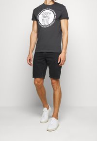 Hombre vestido con una camiseta gráfica negra "Deep Dive", pantalones cortos de mezclilla negros y zapatillas blancas de pie contra un fondo liso.