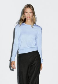 Giovane donna con lunghi capelli biondi che indossa un maglione a maniche lunghe azzurro chiaro e pantaloni neri, in piedi contro uno sfondo bianco semplice.