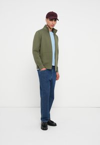 Ελιά πράσινη ζακέτα με φερμουάρ και ψηλό γιακά, μακριά μανίκια; ανοιχτό μπλε μπλούζα από κάτω; κλασικά μπλε τζιν; σκούρα παπούτσια; μπορντό καπέλο.