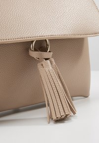 Borsa in pelle beige texture con chiusura a pattina, accentata da un anello metallico e una lunga frangia di nappina attaccata sul davanti.