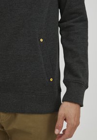 Sudadera gris oscura con dobladillo acanalado, puños en las muñecas y un bolsillo lateral discreto con acentos de botón amarillo. Tejido suave y texturizado.
