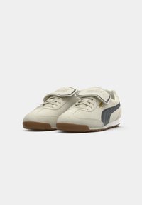 Sko med en blød hvid ruskindsøverdel, mørkegrå Puma-stribe og beige gummisål; har velcrobånd og snørelukning.