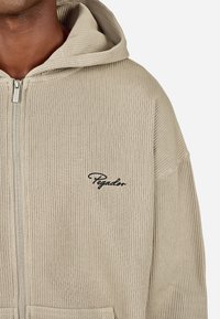 Beige ribbet zip-up hette laget av strukturert stoff, med en frontlomme og brodert "Pegador"-logo på venstre side.
