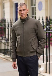 Braune Bomberjacke mit hohem Kragen, zwei Seitentaschen und geripptem Saum, getragen über dunklen Hosen. Verfügt über eine glatte, matte Textur.