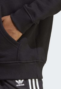 Černá mikina se žebrovanými manžetami, boční kapsou a jemnou texturou. Na černých kalhotách je bílé branding Adidas podél lemu.