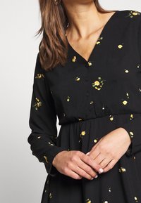 Schwarzes Kleid mit langen Ärmeln, das über einen tiefen V-Ausschnitt und Knöpfe verfügt. Verziert mit verstreuten gelben Blumenmustern und einer gerafften Taille.