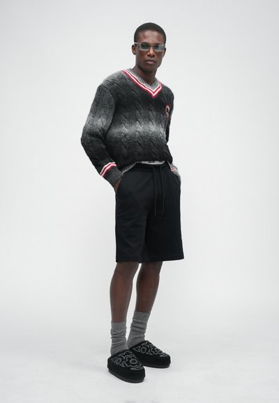 Grauer kabelgestrickter Pullover mit rot-weißen Akzenten, schwarze Shorts, graue Socken und schwarze Hausschuhe mit floralen Mustern.