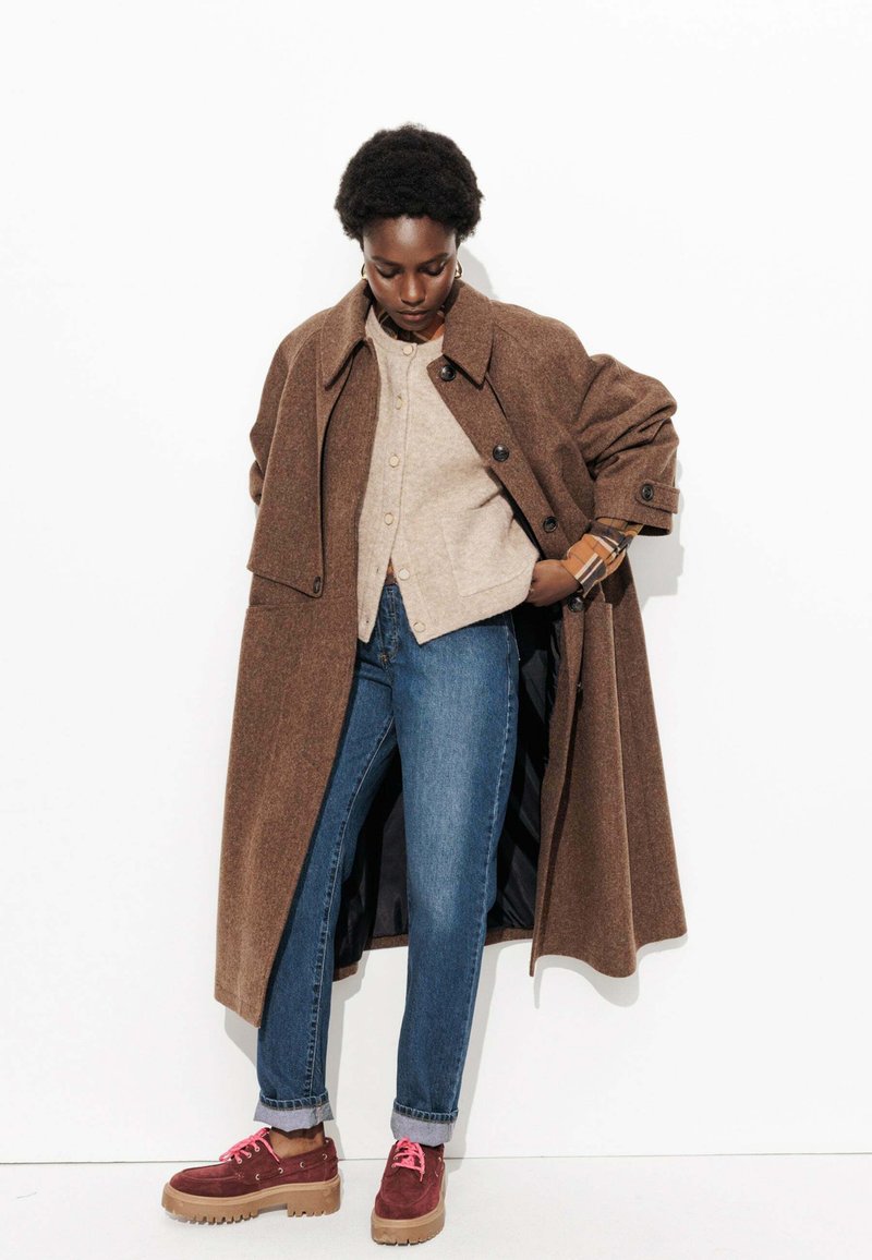 Manteau long en mélange de laine marron avec de gros boutons, chemise beige à boutons, jeans bleus et chaussures en daim bordeaux avec des lacets roses.