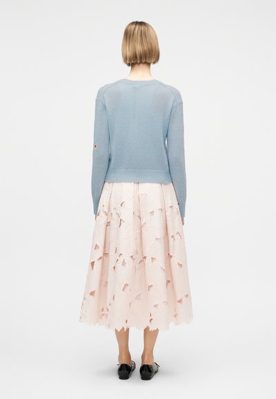 Γυναίκα με κοντά ξανθά μαλλιά, που φοράει γαλάζιο πουλόβερ, απαλό ροζ μίντι φούστα με κοψίματα και μαύρα επίπεδα παπούτσια, στεκόμενη με την πλάτη προς το μέρος μας.