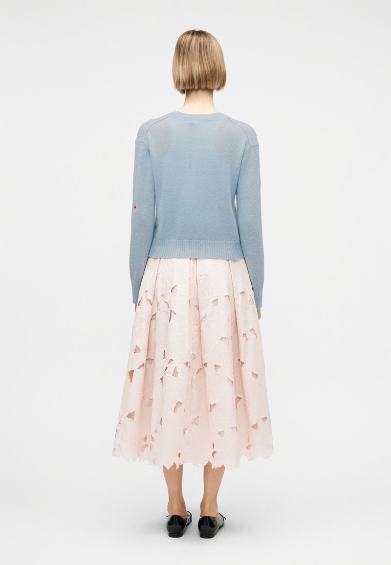 Donna con capelli corti biondi che indossa un maglione azzurro chiaro, una gonna midi rosa pallido con dettagli ritagliati e scarpe basse nere, in piedi di spalle.