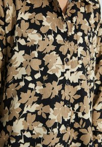 Primo piano di una camicia bottonata con motivi floreali beige e bianchi su un tessuto nero strutturato.