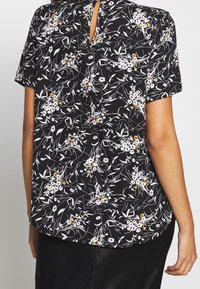Chemise noire à manches courtes avec un motif floral comprenant des fleurs blanches et jaunes, une ouverture en trou de serrure dans le dos, et un ourlet incurvé.
